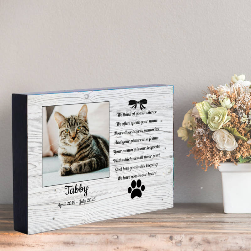 Memorial pet photo frame with a cat's picture and text on a wooden surface with flowers in the background.