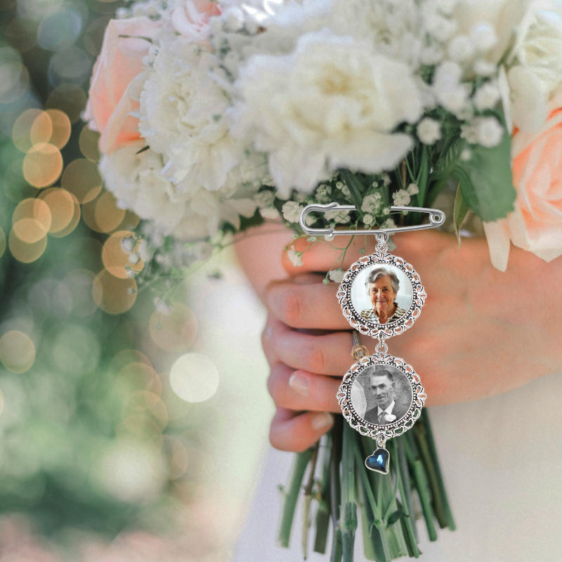 A silver safety pin brooch with two oval portrait frames and a small blue heart charm, held against a bouquet of white and peach flowers. Hand made in Ireland with fast delivery nationwide
