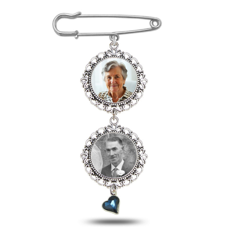 A silver-toned safety pin brooch featuring two circular photo frames with ornate filigree borders, one showing a color portrait of a woman and the other a black-and-white portrait of a man, with a dangling blue heart charm at the bottom. "Something blue" for your wedding