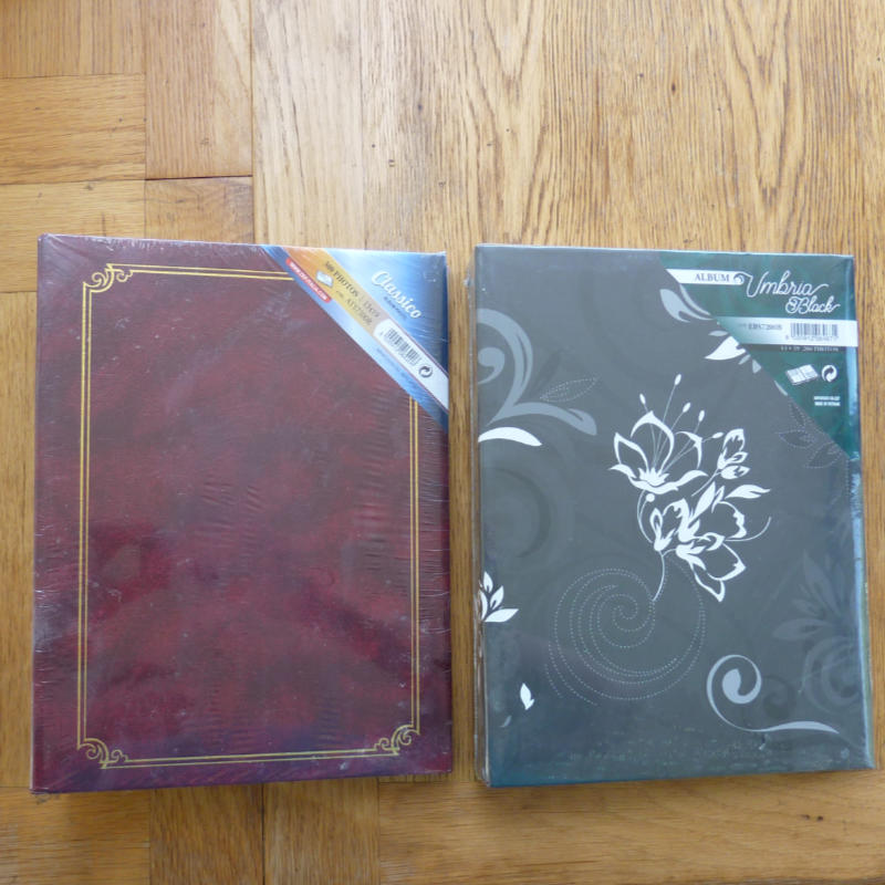 Two decorative photo albums placed side by side on a wooden floor. The left one is dark burgundy with a gold border design, and the right one is dark gray with white floral and swirl patterns.