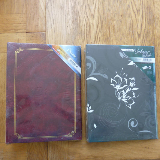 Two decorative photo albums placed side by side on a wooden floor. The left one is dark burgundy with a gold border design, and the right one is dark gray with white floral and swirl patterns.