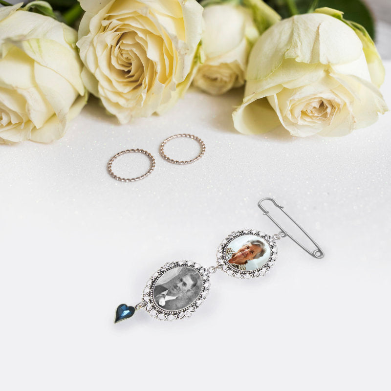 A silver-toned photo keepsake pin brooch with two circular personalised photos, a dangling heart-shaped blue rhinestone,  styled on a white surface with cream roses in the background.