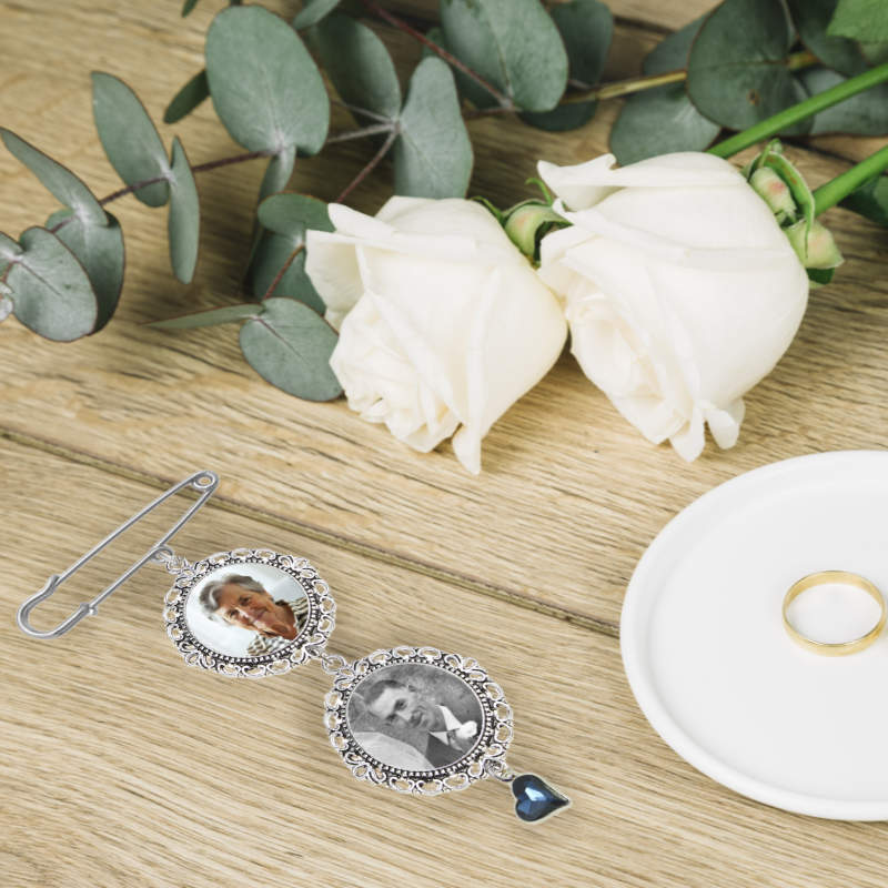 A silver-toned photo keepsake pin with two circular personalised photos and a dangling dark blue heart charm, styled on a wooden surface with white roses, eucalyptus leaves, and a gold ring.