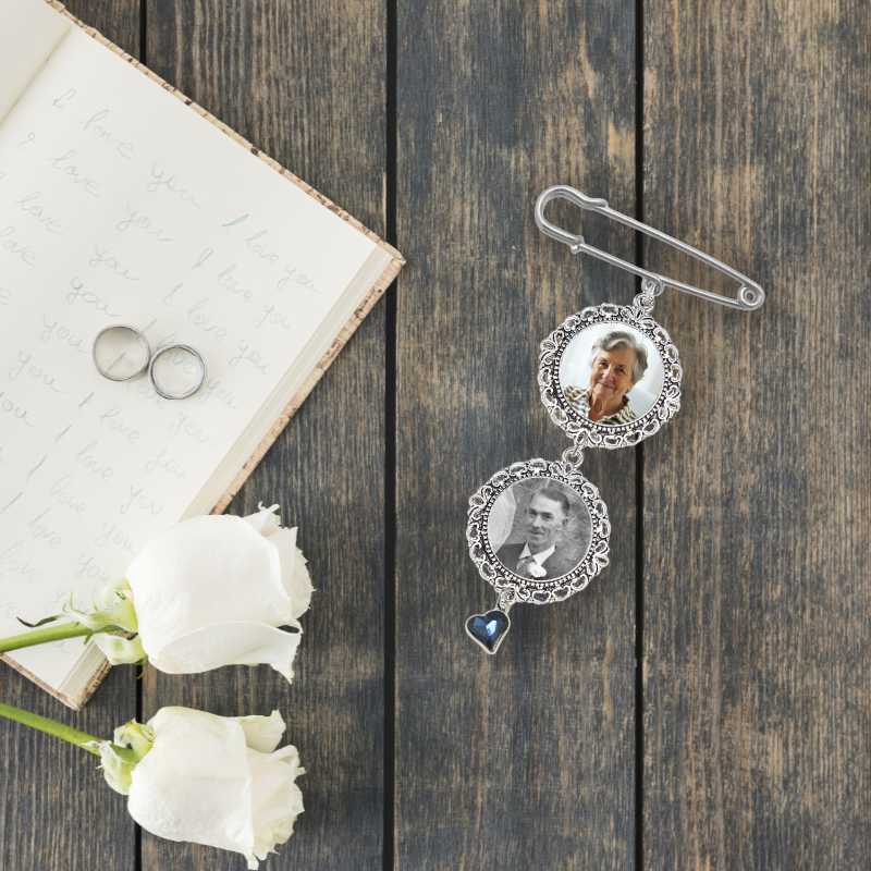 A silver-toned safety pin brooch with two ornate circular photo frames and a dangling heart charm, styled on a dark wood surface beside an open handwritten book, two rings, and white roses.