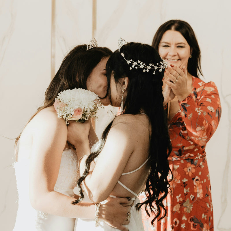 Two women in wedding dresses embracing, with a third woman in a floral dress standing behind them.