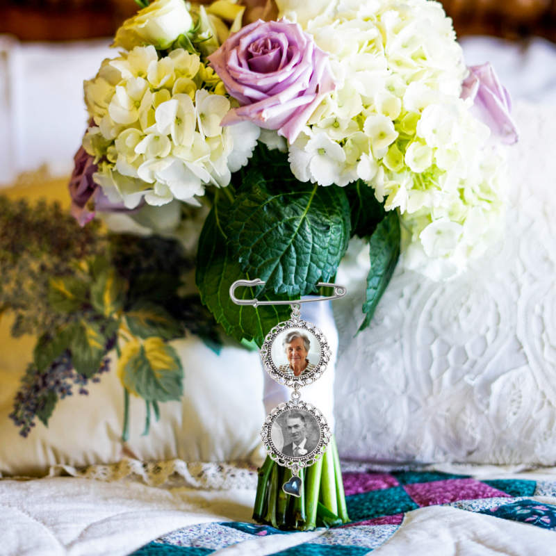 A close-up of a bouquet of white and lavender hydrangeas with a purple rose, featuring a silver brooch holding two circular photo frames with a rhinestone blue heart.